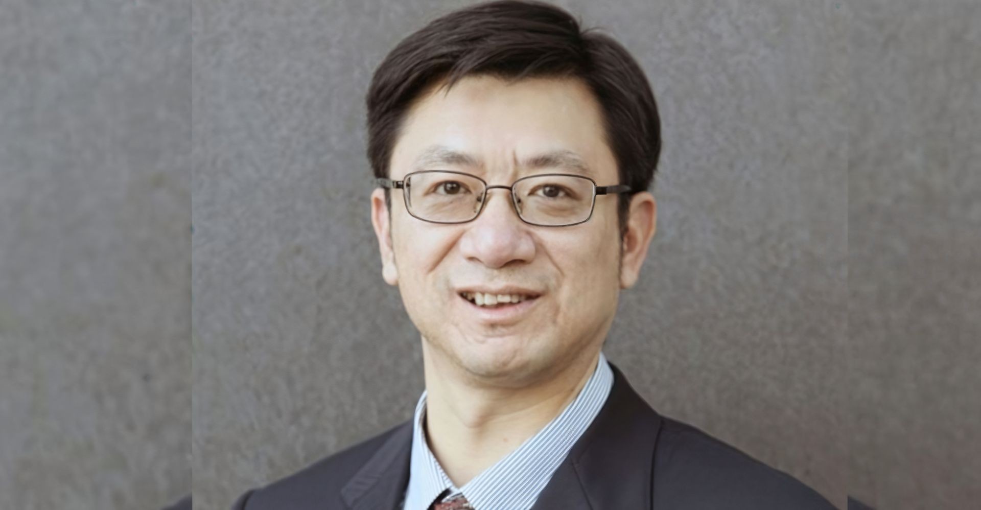 Man with dark hair wearing a suit and tie, standing against a gray textured background.